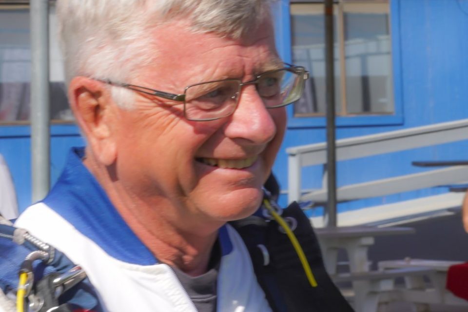 An older man with gray hair and glasses is smiling outdoors. He is wearing a skydiving suit with badges on it. A blue building and a canopy are visible in the background.