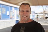 A smiling man in a black T-shirt stands outdoors under a canopy. The background shows blurred structures with blue and white walls.