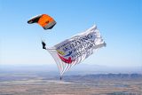 A skydiver in mid-air holds a large banner with the text "International Skydiving Museum & Hall of Fame." The parachute is orange and white, and the landscape below is a vast expanse of mountains and plains under a clear blue sky.