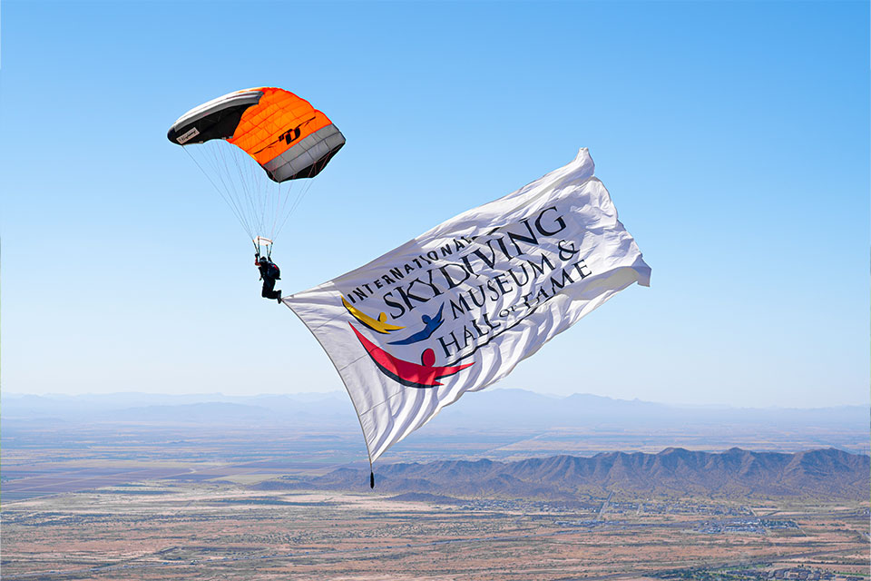 A skydiver in mid-air holds a large banner with the text "International Skydiving Museum & Hall of Fame." The parachute is orange and white, and the landscape below is a vast expanse of mountains and plains under a clear blue sky.