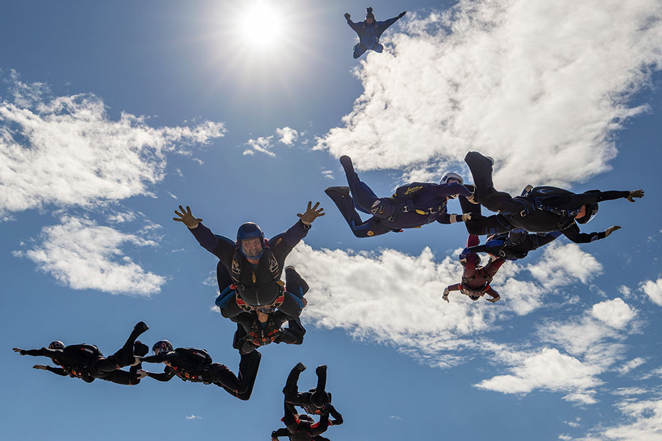 Group of skydivers in mid-air, performing a formation under a bright sun. They wear helmets and suits against a backdrop of a clear blue sky with scattered clouds.