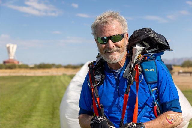 A smiling man with gray hair stands in a grassy field, wearing a blue shirt, sunglasses, and a parachute. The sky is clear, and there's a structure in the background. He's holding his gear and looks content.