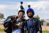 Two smiling skydivers in gear stand close together outdoors. Each has a helmet with cameras attached. The person on the left carries a parachute. The sky is clear, and there are structures and trees in the background.