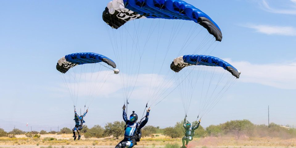 Three skydivers in blue gear and parachutes land gracefully on a grassy field under a clear blue sky. Sparse desert vegetation is visible in the background.