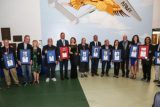 A group of people stands in a line holding framed awards. They are in a hall with a mural featuring an eagle and the word "Army" on the wall behind them. Everyone is dressed formally and smiling for the photo.