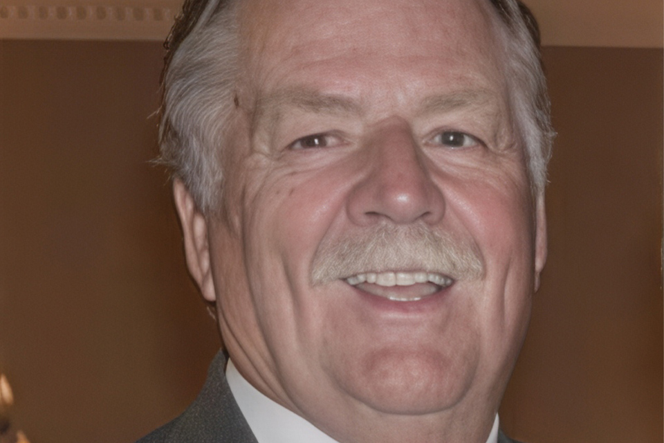 A smiling older man with gray hair and a mustache is wearing a dark suit and white shirt. The background is softly blurred, focusing attention on the man's friendly expression.