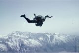 A skydiver in mid-air against a backdrop of snow-capped mountains. The person is wearing a helmet and jumpsuit, with arms and legs extended in a free-fall position. The sky is clear, enhancing the view of the rugged peaks below.