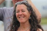 A woman with long, curly hair smiles outdoors. She is wearing hoop earrings and sunglasses on her head. The background is blurred, showing a casually dressed person behind her.