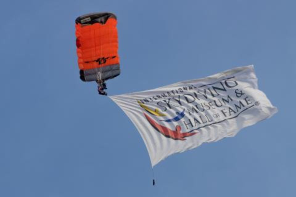 A parachutist descends with an orange and gray parachute, holding a large banner that reads "International Skydiving Museum & Hall of Fame" against a clear blue sky.