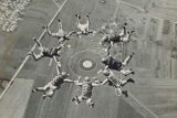 Seven parachutists form a circular formation mid-air over a rural landscape, with fields and a runway visible below. They appear to be synchronized as they skydive on a clear day.