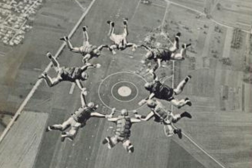 Seven parachutists form a circular formation mid-air over a rural landscape, with fields and a runway visible below. They appear to be synchronized as they skydive on a clear day.