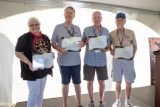 Four older adults stand on a stage, each holding a certificate or plaque. They are smiling and wearing casual clothes and medals around their necks. A white tent serves as the backdrop.