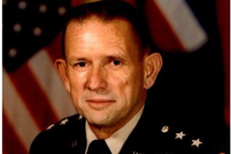 A person in a military uniform poses in front of a blurred background with an American flag. They have short hair and a neutral expression.