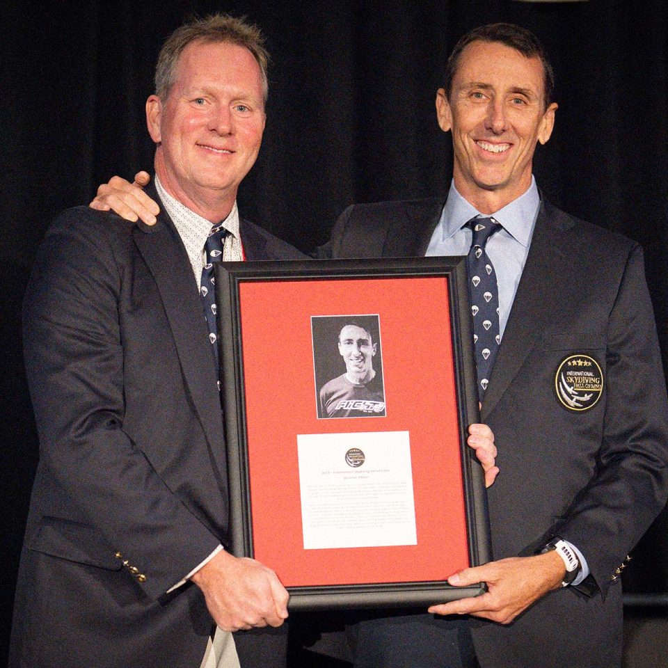 Two men in suits smiling and holding a framed award with a photograph and text on it. One man wears a badge on his jacket. They stand against a black backdrop.