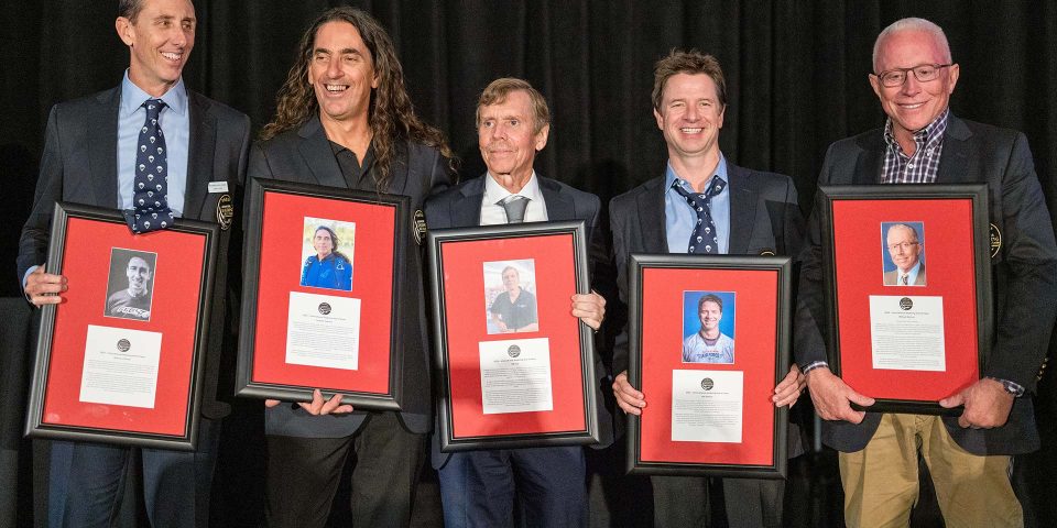 Five men stand in front of a dark curtain, each holding a framed plaque with a portrait and text. They are dressed in suits, smiling, and appear to be at an awards or recognition event.