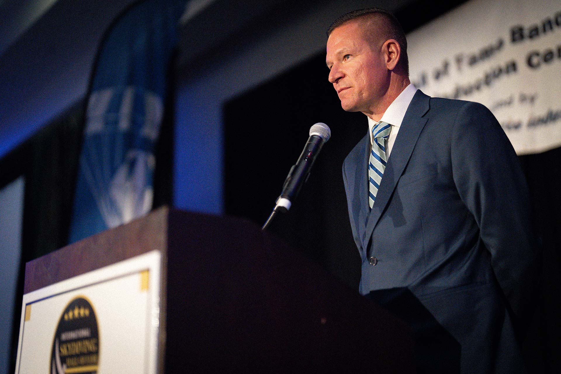 A man in a blue suit and striped tie stands at a podium with a microphone, delivering a speech. The background features a banner with partially visible text and a dark curtain.