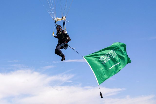 A skydiver is gracefully descending against a clear blue sky, carrying the national flag of Saudi Arabia. The vibrant green flag with white Arabic script and a sword flutters in the wind, creating a striking visual.