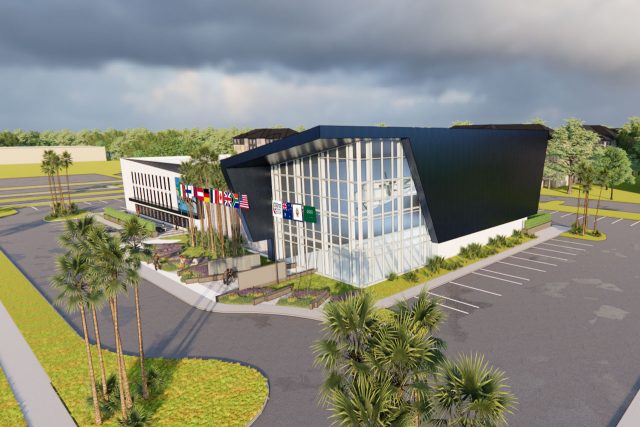 A modern building with angular architecture featuring a glass facade, surrounded by a parking lot and palm trees. Multiple national flags are displayed at the entrance. The sky is overcast with dark clouds.