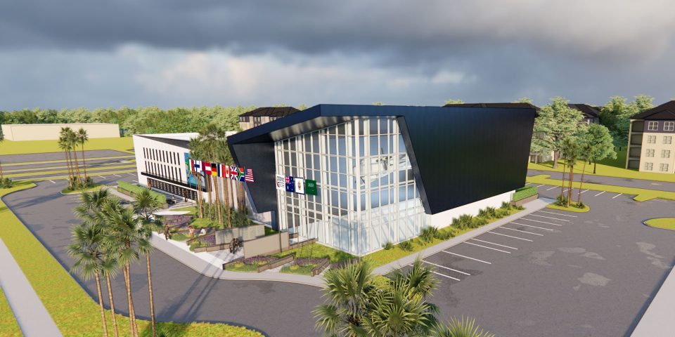 Modern office building with a sleek black facade and large windows, surrounded by parking spaces and landscaping with palm trees. Flags from various countries are displayed on the side. Cloudy sky in the background.