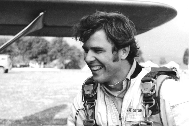 A person wearing a skydiving suit and gear is smiling and standing outdoors near an aircraft wing. The scene is in black and white, with trees and a field in the background.