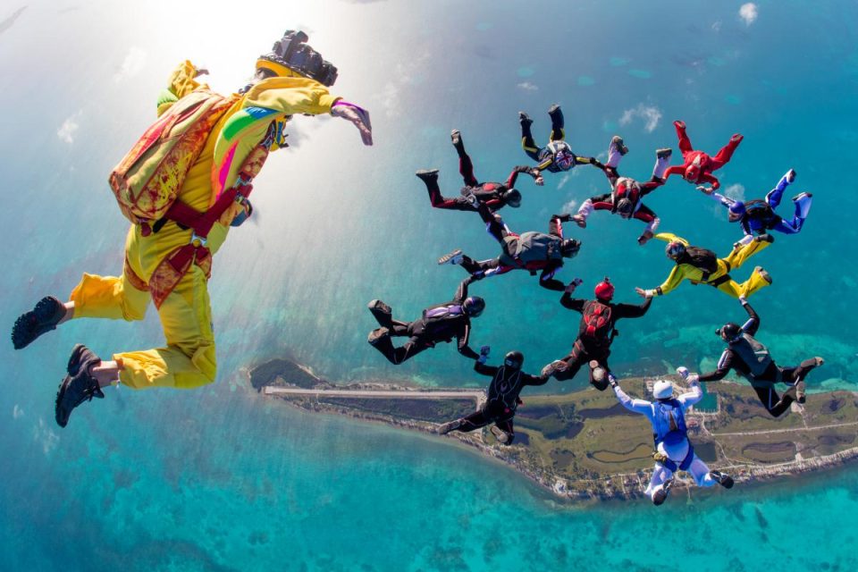 A group of skydivers in colorful outfits form a circle formation high above a coastline with vibrant blue waters. One additional skydiver in yellow seems to be capturing the scene. The sun shines brightly over the horizon.