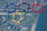 Skydivers form the Olympic rings in mid-air, each wearing jumpsuits matching the ring colors: blue, yellow, black, green, and red. The cityscape below includes a stadium and other buildings near a body of water.