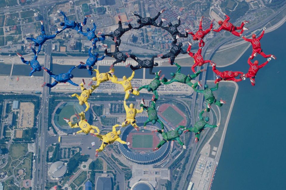 Skydivers form the Olympic rings in mid-air, each wearing jumpsuits matching the ring colors: blue, yellow, black, green, and red. The cityscape below includes a stadium and other buildings near a body of water.