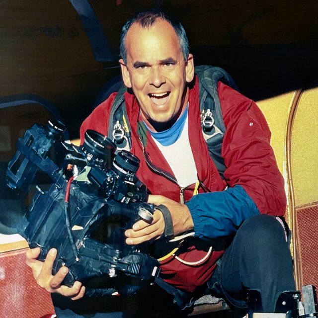 A person smiling while sitting in an aircraft, holding a complex camera rig with multiple lenses. They are wearing a red jacket and a backpack, suggesting they are involved in aerial photography or filming.