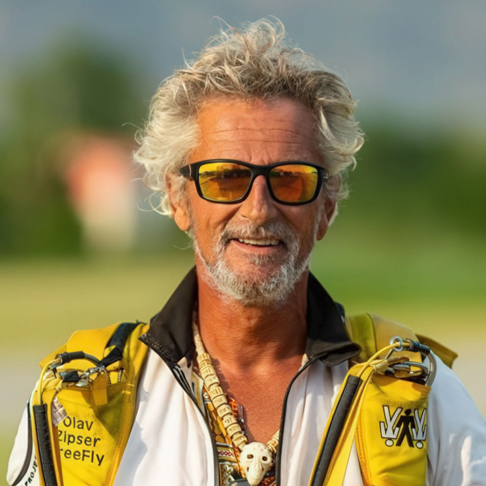 Man with white hair and glasses wearing a yellow and white jacket, smiling outdoors.