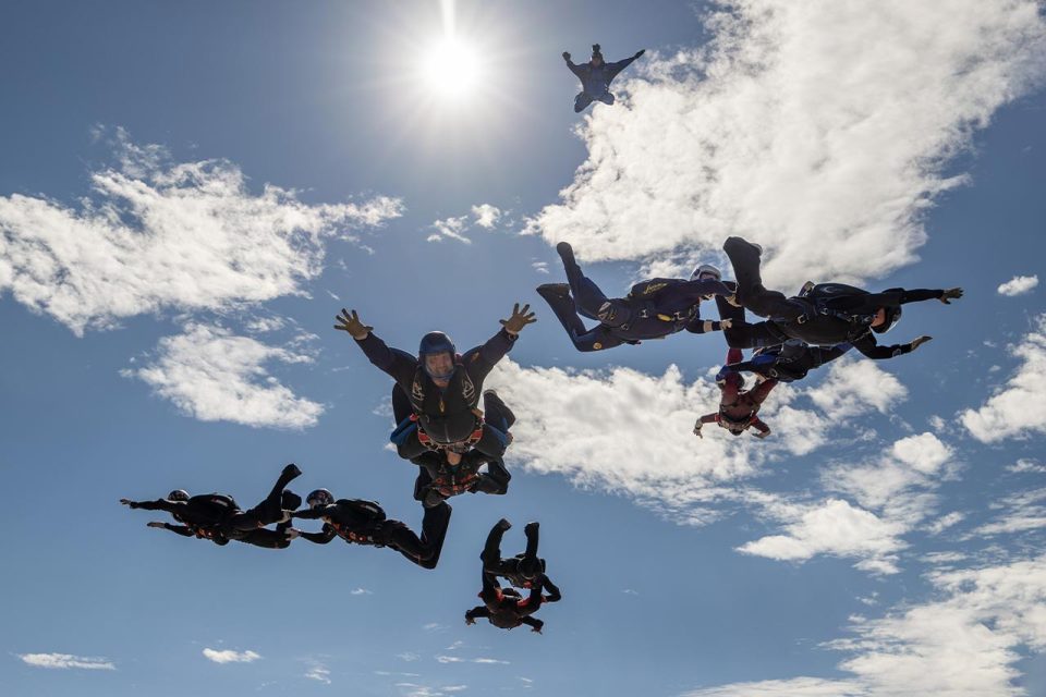 A group of skydivers in freefall, wearing helmets and jumpsuits, forming a pattern against a bright blue sky with scattered clouds. The sun shines brightly, adding dramatic lighting to the scene.