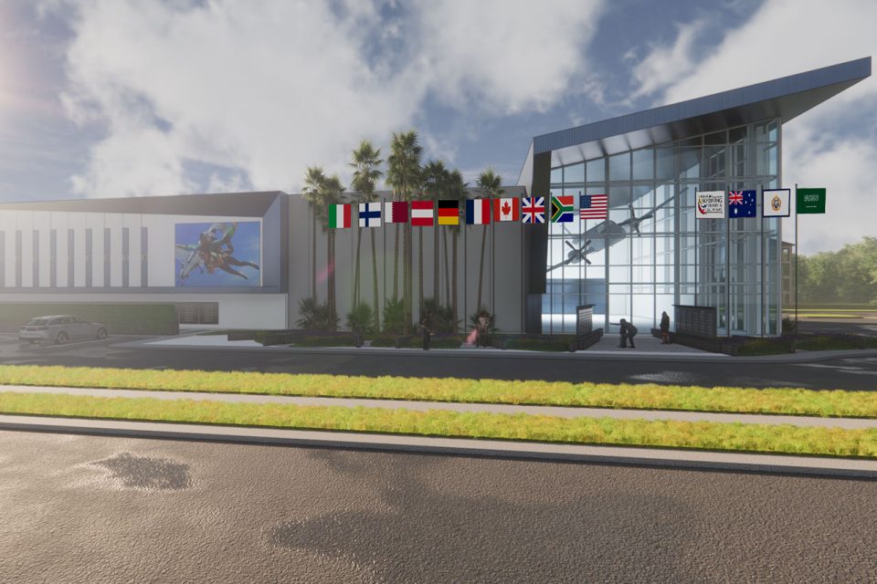 Modern building with a glass entrance and multiple international flags, including Germany and Canada, displayed above. Artwork of an insect on the exterior wall. Few people walking outside on a sunny day.