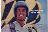 A man wearing a helmet and skydiving suit smiles in front of a large, unfurled parachute. The text on the image reads "Championship in the Sky." His suit has patches, including an American flag.