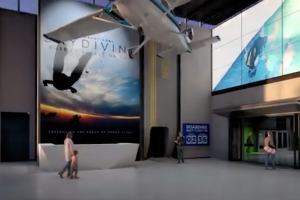 A modern airport lobby with a skydiving theme. Large posters and screens display images of skydivers. A small plane model hangs from the ceiling. People are walking and standing near a boarding gate sign displaying flight times.