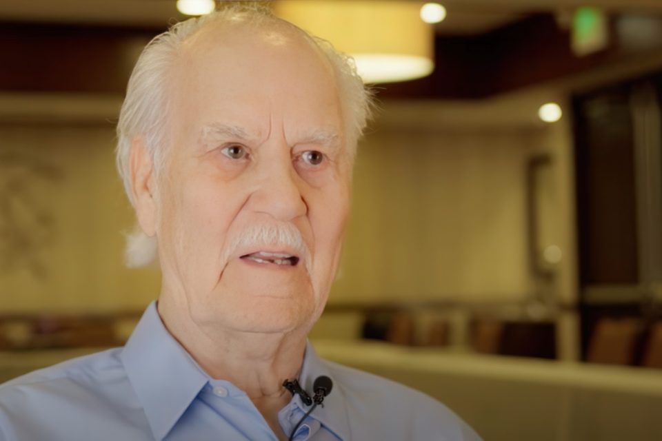 An older man with white hair and a mustache is wearing a light blue shirt and a microphone. He is indoors, with a softly lit background. The setting appears professional, possibly a conference or an interview.