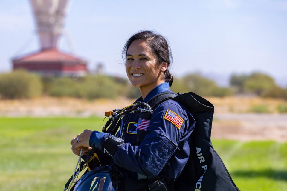 A person in a blue Air Force jumpsuit stands outdoors, holding a parachute. They are smiling with a structure in the distant background, surrounded by greenery and a clear sky.