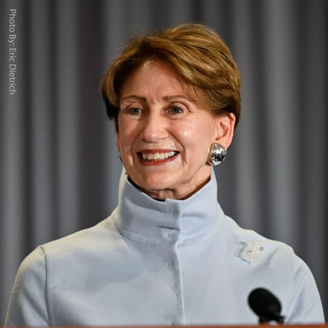 A person with short hair smiles while wearing a light blue blazer with a high collar. They have silver earrings and stand in front of a gray curtain. A microphone is visible in the foreground.