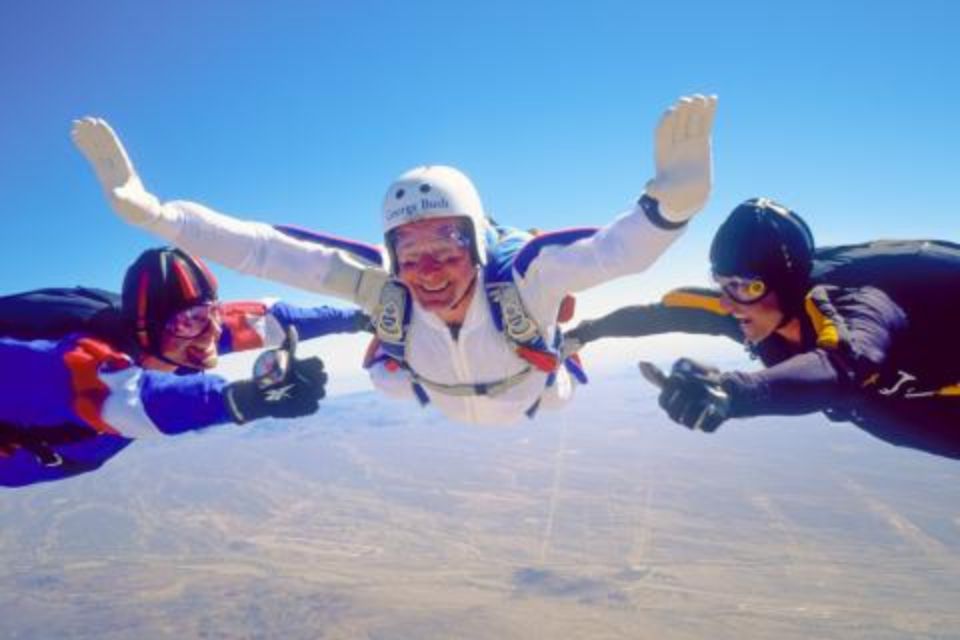 Three skydivers in free fall with clear blue skies in the background. The central skydiver is wearing a white jumpsuit and helmet, while the other two are in multicolored outfits. They are smiling and giving thumbs-up gestures.