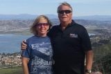 A woman and a man stand side by side in front of a scenic landscape with a lake, hills, and a town below. Both are wearing sunglasses and smiling, with the man wearing a dark shirt and the woman a Led Zeppelin T-shirt.