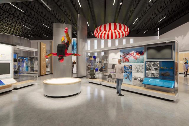 A museum exhibit showcasing space exploration is displayed. A mannequin in a spacesuit hangs in a dynamic pose, and visitors examine various artifacts and informational panels. The ceiling features a red and white striped parachute.