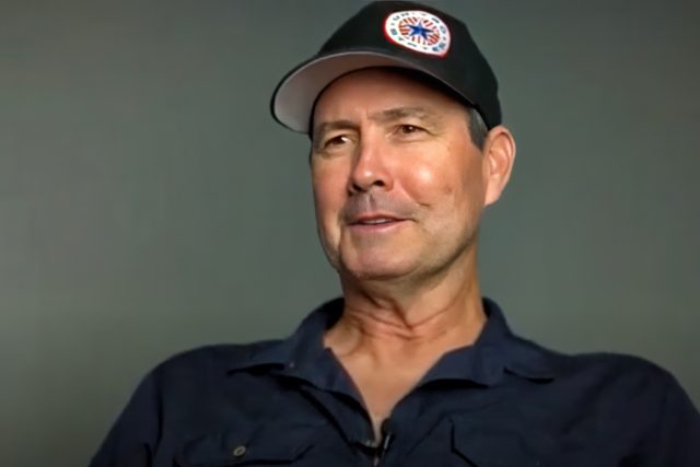 A man wearing a black cap with a colorful circular logo and a dark button-up shirt smiles slightly against a gray background.