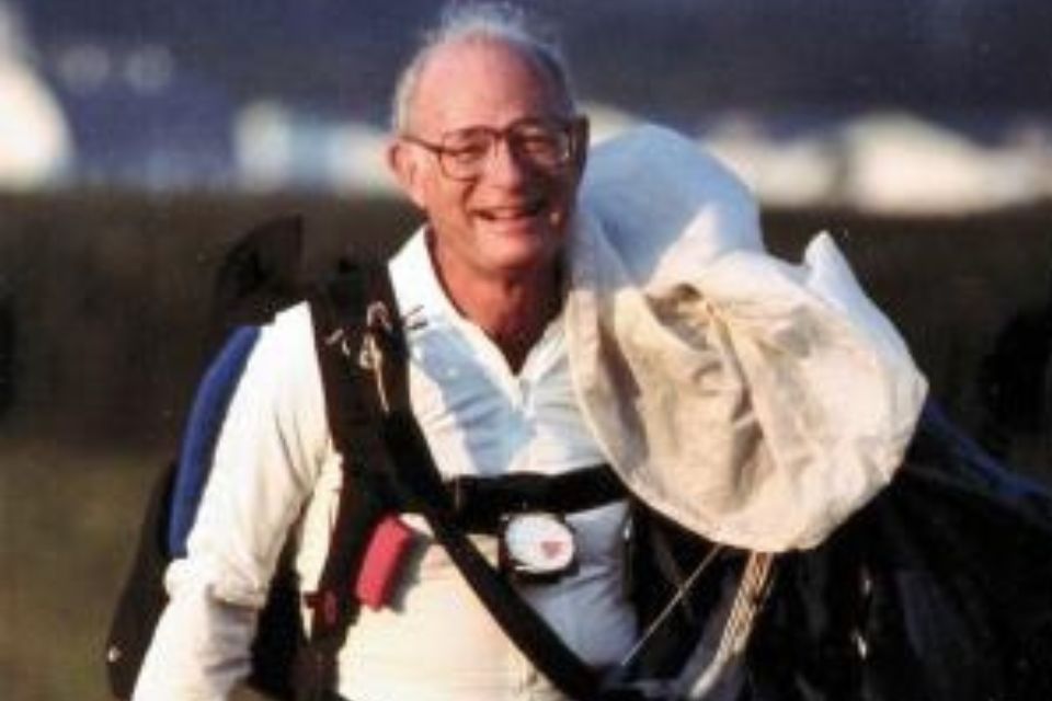 An older man smiles warmly as he carries a parachute and wears a skydiving harness. He's outdoors, with blurred greenery and structures in the background.