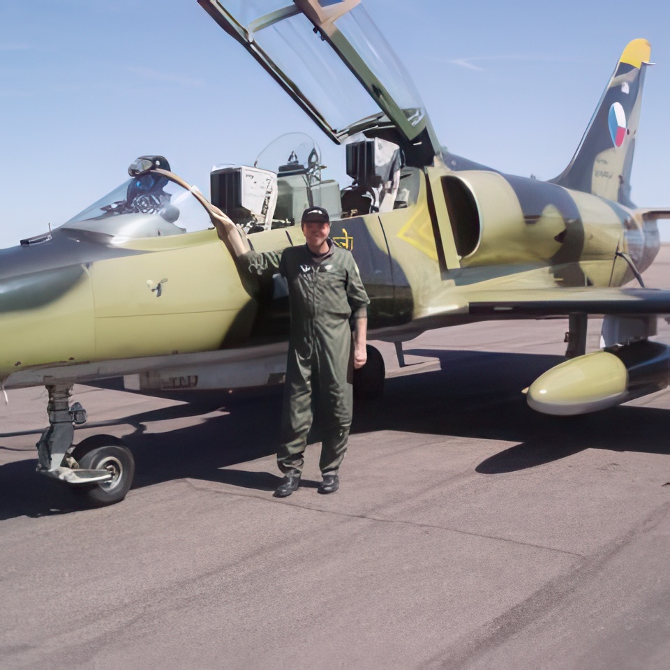 A person in a green flight suit stands beside a sleek, camouflaged military jet on a runway, with the cockpit open. The jet has a distinctive tail emblem and the sky is clear and blue.