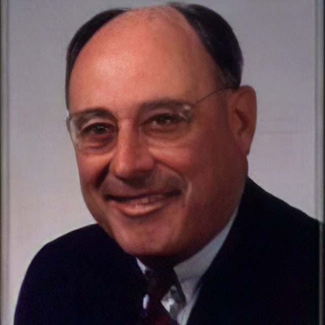 A man with glasses, short dark hair, and a receding hairline smiles in a formal portrait. He's wearing a dark suit jacket, white shirt, and red tie against a plain background.