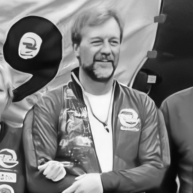 A person with a distinctive beard and mustache is smiling while wearing a jacket with logos. The background features a fabric backdrop and a partial figure on the right. The image is in black and white.