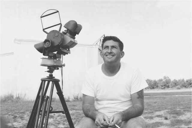 A man in a white t-shirt sits outdoors next to a telescope on a tripod. He is smiling and holding an object in his hand. The background features a structure and open sky.