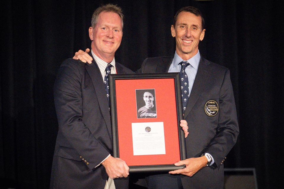 Two men in suits smiling and holding an award plaque with a black-and-white photo and text. The man on the right wears a badge on his jacket. The background is dark, suggesting an indoor event or ceremony.