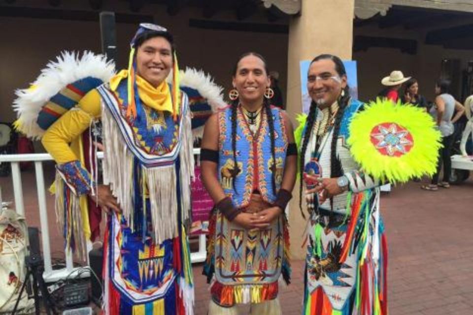 Three people in colorful traditional Native American regalia stand together, smiling at an outdoor event. The regalia features intricate beadwork, fringes, and vibrant colors. They hold decorative fans and wear feathered accessories.