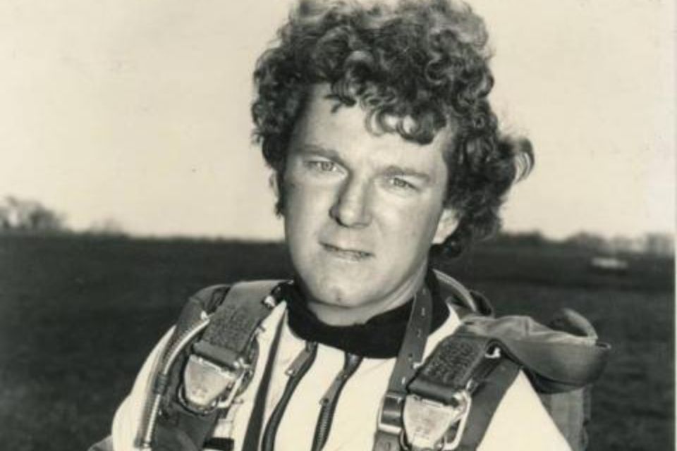 A person with curly hair is wearing a parachute harness and looking at the camera. The background is an open field on a clear day. The image is in black and white.