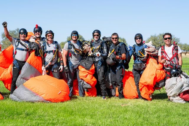 A group of eight skydivers standing on grass, smiling and holding bright orange parachutes. They are wearing helmets, harnesses, and jumpsuits. The sky is clear, and the background has trees and open space.