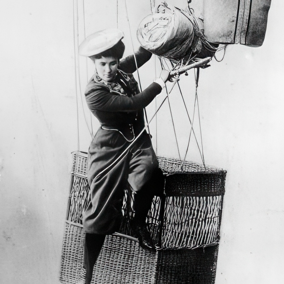 A person dressed in early 20th-century attire hangs onto a wicker basket of a hot air balloon. They wear a hat and boots, gripping ropes attached to the balloon's fabric, with luggage tied above. The image is in black and white.